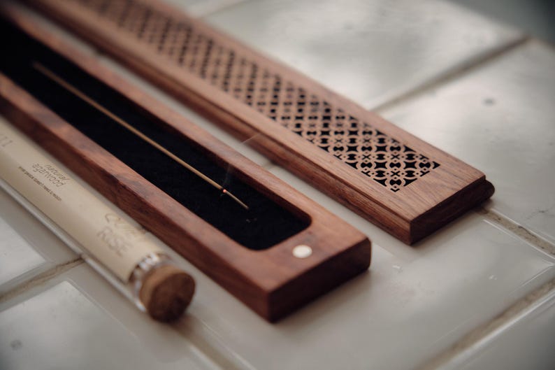 May include: A wooden incense holder with a decorative, geometric pattern on the lid. The holder is open, revealing a lit incense stick. A glass tube with text is in the foreground, and the holder is on a tiled surface.