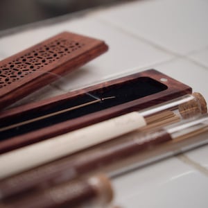 May include: A wooden incense holder with a decorative carved lid, alongside several glass tubes containing incense sticks. The holder is open, revealing a lit incense stick. The tubes have cork stoppers and white labels.