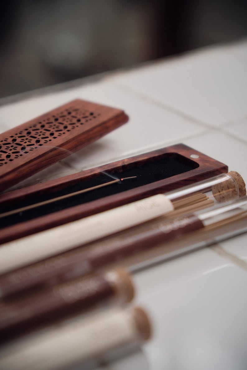 May include: A wooden incense holder with a decorative carved lid, alongside several glass tubes containing incense sticks. The holder is open, revealing a lit incense stick. The tubes have cork stoppers and white labels.
