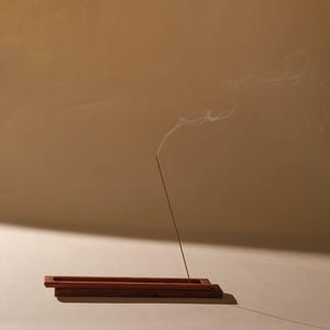 May include: A wooden incense holder with a burning incense stick, emitting a wisp of smoke. The holder is rectangular with a groove to catch ash. The background is a warm, neutral tone, creating a serene and minimalist aesthetic.