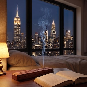 May include: A nighttime cityscape view through a large window, featuring the Empire State Building and other skyscrapers. A lit lamp, a wooden incense holder, and an open book sit on a wooden table. Wisps of smoke rise from the incense holder.