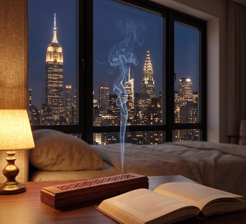 May include: A nighttime cityscape view through a large window, featuring the Empire State Building and other skyscrapers. A lit lamp, a wooden incense holder, and an open book sit on a wooden table. Wisps of smoke rise from the incense holder.