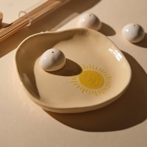 May include: A ceramic incense holder with a yellow sun design on a cream-colored plate. Three white incense holders are next to the plate. A glass tube with incense sticks is in the background.