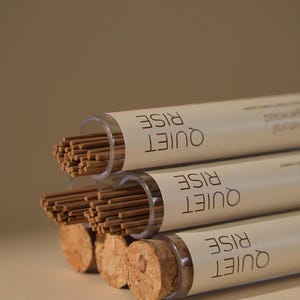 May include: Three tubes of incense sticks with cork stoppers. The tubes are labeled "QUIET RISE" in a stacked format. The incense sticks are brown, and the tubes are clear. The background is a solid, neutral color.