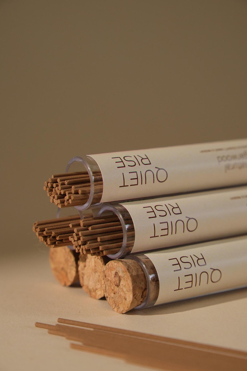May include: Three tubes of incense sticks with cork stoppers. The tubes are labeled "QUIET RISE" in a stacked format. The incense sticks are brown, and the tubes are clear. The background is a solid, neutral color.