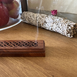 May include: A wooden incense burner with a decorative cut-out design sits on a wooden surface. A bundle of white sage tied with twine and a dried rose are in the background. Smoke rises from the sage. Apples are in a glass bowl.