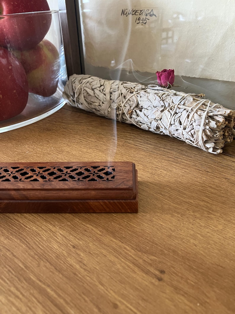 May include: A wooden incense burner with a decorative cut-out design sits on a wooden surface. A bundle of white sage tied with twine and a dried rose are in the background. Smoke rises from the sage. Apples are in a glass bowl.