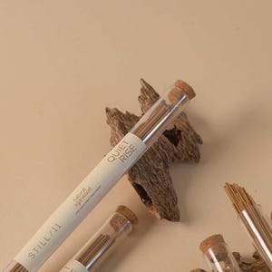 May include: Incense sticks in clear tubes with cork stoppers, labeled "QUIET RISE" and "STILL/11". The sticks are brown, and the tubes rest on pieces of wood against a beige background. The text "natural agarwood" is also visible.