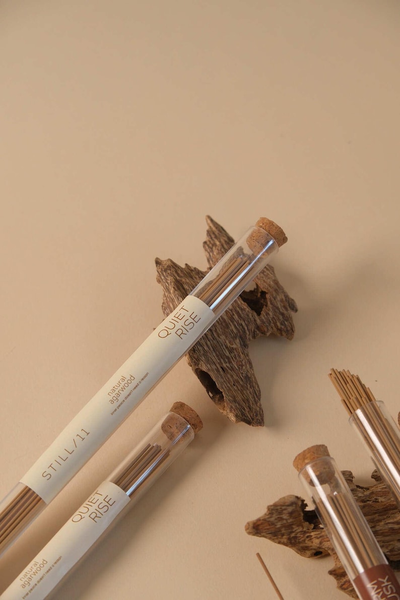 May include: Incense sticks in clear tubes with cork stoppers, labeled "QUIET RISE" and "STILL/11". The sticks are brown, and the tubes rest on pieces of wood against a beige background. The text "natural agarwood" is also visible.