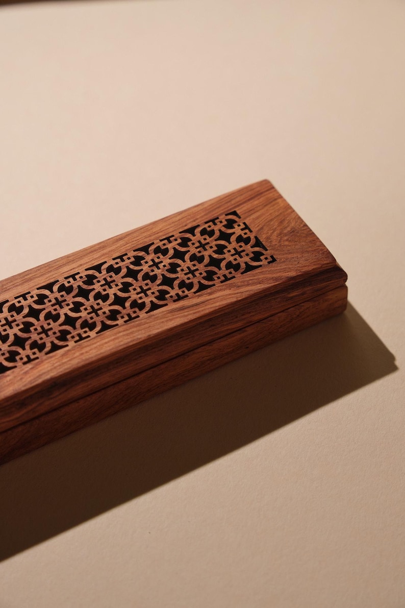 May include: A rectangular wooden box with a decorative geometric pattern carved into the lid. The box is made of a dark brown wood and has a black interior. The design is symmetrical and adds a touch of elegance.