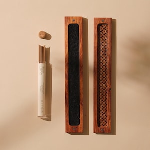 May include: An overhead shot shows a set of incense products. A clear tube of incense sticks with the text "QUIET RISE" and "STILL 11" is next to a wooden incense holder with a black felt interior. A second wooden holder with a decorative cutout design is also present.