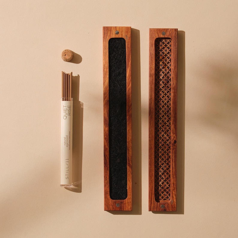 May include: An overhead shot shows a set of incense products. A clear tube of incense sticks with the text "QUIET RISE" and "STILL 11" is next to a wooden incense holder with a black felt interior. A second wooden holder with a decorative cutout design is also present.