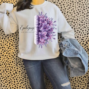 Op de afbeelding: Witte sweatshirt met een bloemenontwerp in paarse en lavendeltinten, met de woorden "Epilepsy Awareness". Het ontwerp bevat een gestileerd brein en bloemen. De sweatshirt heeft lange mouwen.