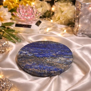 Puede incluir: Un posavasos redondo de piedra de lapislázuli pulido, de color azul intenso con motas doradas. El posavasos se muestra sobre una superficie de satén blanco, con flores y luces de hadas en el fondo.