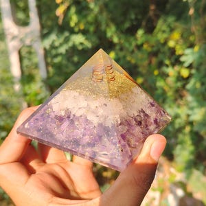 Könnte beinhalten: Handgefertigte pyramidenförmige Dekoration mit Schichten aus lila Amethyst, weißen Kristallen und goldenem Glitzer. Der obere Bereich hat eine goldene Spule. Das Objekt wird in einer Hand vor einem verschwommenen grünen Hintergrund gehalten.