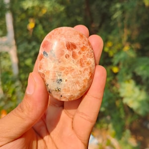 May include: A polished, oval-shaped sunstone with a speckled, orange and cream colour pattern. The stone is held in a hand, with a blurred green background. The stone is approximately 5 cm in length.