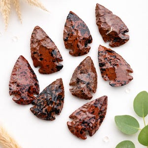 May include: A collection of eight reddish-brown obsidian arrowheads with black veining. The arrowheads are arranged on a white surface, with decorative elements of wheat stalks and green leaves.