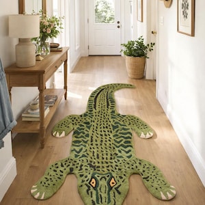May include: A green and black alligator-shaped rug with a textured pattern on a wooden floor in a hallway. The rug has detailed eyes, teeth, and feet. The hallway has white walls, a wooden console table, and a mirror. The rug measures approximately 200 cm in length.