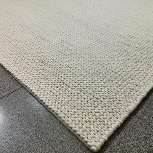 May include: A close-up of a cream-colored, hand-knitted rug. The rug has a thick, textured appearance with visible stitches. The rug is placed on a gray tiled floor, with the corner of the rug in the foreground.