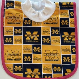 Könnte beinhalten: Ein Baby-Lätzchen mit einem sich wiederholenden Muster des Logos der University of Michigan in gelben und marineblauen Quadraten. Es hat ein weißes Halsband und eine rosa Umrandung. Die Texte "MICHIGAN" und "THE UNIVERSITY OF MICHIGAN" sind sichtbar.