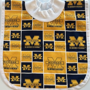 Könnte beinhalten: Ein Baby-Lätzchen mit einem sich wiederholenden Muster aus marineblauen und gelben Quadraten mit dem Text "MICHIGAN" und dem Logo der University of Michigan. Es hat eine weiße Einfassung und eine weiße Halsöffnung.