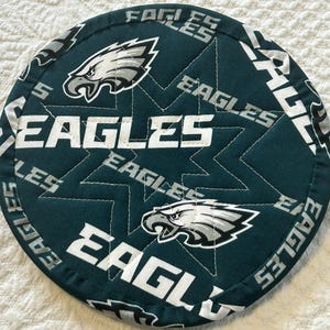 May include: A round, quilted fabric pot holder in dark green featuring a repeating pattern of the Philadelphia Eagles football team logo and the word EAGLES in white and grey text, with a star-shaped stitching pattern across the surface.