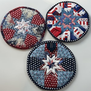 Puede incluir: Tres salvamanteles redondos acolchados patrióticos. Cada salvamanteles presenta un diseño de estrella con telas rojas, blancas y azules. Los salvamanteles tienen un borde azul oscuro con detalles de estrellas blancas. Los salvamanteles son probablemente hechos a mano.