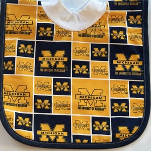 Könnte beinhalten: Ein Baby-Lätzchen mit einem sich wiederholenden Muster des Logos der University of Michigan in Marineblau und Maisgelb. Das Lätzchen hat einen weißen Kragen und eine marineblaue Bordüre.