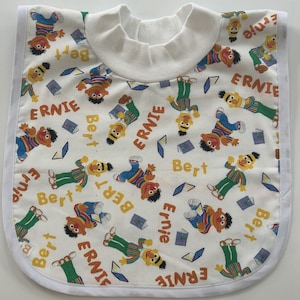 Könnte beinhalten: Weißes Baby-Lätzchen mit einem Muster aus Ernie- und Bert-Figuren, Büchern und Laptops. Das Lätzchen hat eine weiße Einfassung und einen weißen Kragen. Die Namen "Ernie" und "Bert" sind aufgedruckt.
