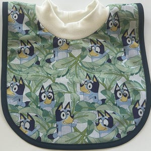 May include: A baby bib featuring a repeating pattern of a cartoon dog character in a jungle-themed design. The bib has a white turtleneck collar and is trimmed with a dark blue border.