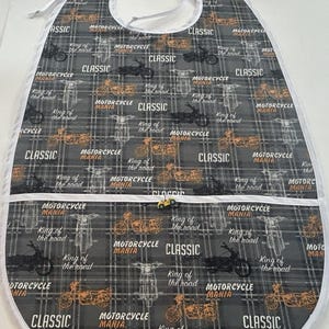 May include: A gray and white plaid baby bib with a motorcycle theme. The bib features a repeating pattern of motorcycles in orange, white, and black, with the words "MOTORCYCLE MANIA" and "CLASSIC". It has a white trim and a pocket.