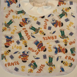 Könnte beinhalten: Weißes Baby-Lätzchen mit einem Muster aus Ernie- und Bert-Figuren, Büchern und den Namen "Ernie" und "Bert" in Gelb. Das Lätzchen hat eine weiße Einfassung und einen weißen Halsbund.