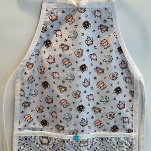 May include: White apron with a light blue background and a pattern of teapots in various colors. The apron has a pocket with a gray and white diamond pattern and a small blue flower. White straps and trim complete the design.