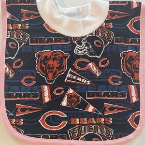 May include: A baby bib featuring a navy blue background with a repeating pattern of Chicago Bears logos, helmets, and bear head graphics in orange and white. The bib has a white collar and a pink trim.