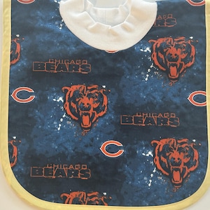 May include: A baby bib featuring a navy blue background with a pattern of orange Chicago Bears logos and text. The bib has a white collar and a yellow trim.