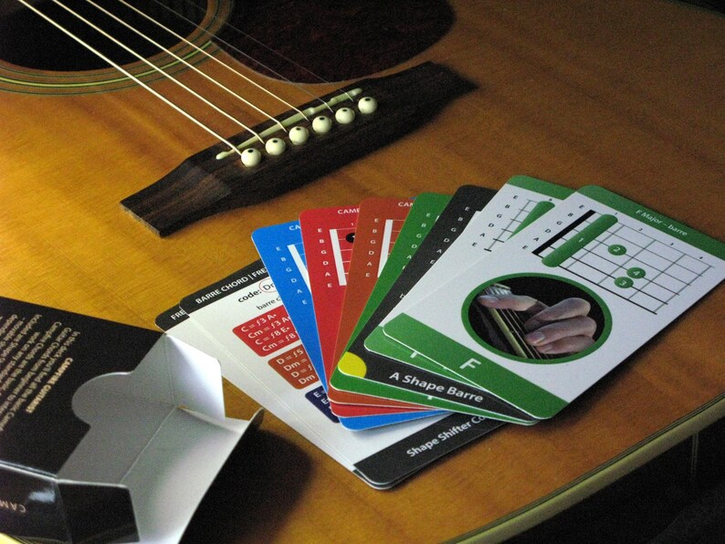 May include: Acoustic guitar with a set of colorful guitar chord cards fanned out on the wooden surface. The cards display chord diagrams and finger positions, with text including "Barre Chord F" and "A Shape Barre". A black box is in the lower left.