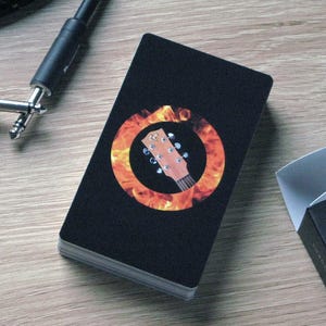 May include: A deck of black Campfire Guitarist playing cards with a guitar headstock graphic inside a flame circle. A black guitar cable and a partially visible black card box are also present on a wooden surface.