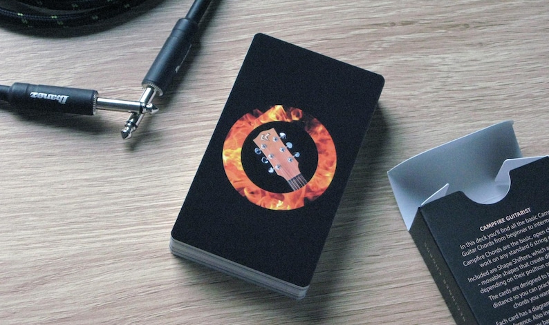 May include: A deck of black Campfire Guitarist playing cards with a guitar headstock graphic inside a flame circle. A black guitar cable and a partially visible black card box are also present on a wooden surface.