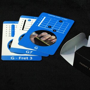 May include: A deck of blue and white guitar chord cards with the text "G", "G7", and "G - Fret 3". The cards show finger placement diagrams. A black box with the text "BEGINNER CHORDS" is partially visible.
