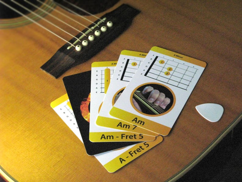 May include: Acoustic guitar with strings and bridge, alongside a collection of guitar chord cards. The cards display chord diagrams for Am, Am7, and A-Fret 5, with a white guitar pick resting nearby. The background is a warm, wooden surface.