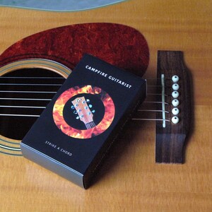 May include: Acoustic guitar with a dark brown soundhole and a red pickguard. A black box with the text "Campfire Guitarist" and "Strike a Chord" is resting on the guitar. The box features a guitar neck graphic.