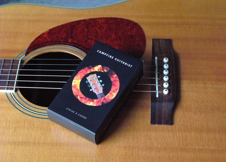 May include: Acoustic guitar with a dark brown soundhole and a red pickguard. A black box with the text "Campfire Guitarist" and "Strike a Chord" is resting on the guitar. The box features a guitar neck graphic.