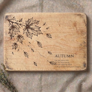 Puede incluir: Una tabla de cortar rectangular de madera con veta natural de color marrón claro. La superficie presenta una ilustración grabada en negro de hojas de arce movidas por el viento, la palabra AUTUMN y el texto: Let the leaves fall, the fire burn warm, and the table be filled with good food and good company.