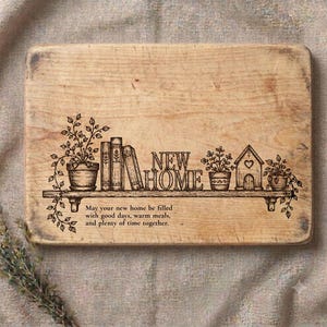 May include: A rectangular light-coloured wooden cutting board featuring a laser-engraved illustration of a shelf with potted plants, books, and a house. The text reads: New Home. May your new home be filled with good days, warm meals, and plenty of time together.