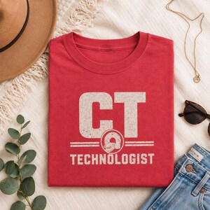 May include: A red crew neck t-shirt featuring the white distressed text CT TECHNOLOGIST with a graphic of a CT scanner machine centered between the words. The design is printed in a bold, block font style on the front of the garment.