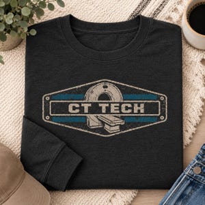 May include: A black long-sleeve t-shirt featuring a distressed graphic of a CT scanner inside a hexagonal frame. The design includes a horizontal blue stripe and the text CT TECH in a bold, white, sans-serif font centered on the chest.