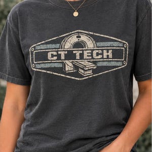 May include: A charcoal gray short-sleeve t-shirt featuring a distressed graphic print of a CT scanner inside a hexagonal frame with the text CT TECH in bold white letters. The design includes muted blue horizontal stripes and a weathered texture.