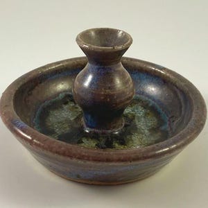 May include: A handmade ceramic candle holder with a dark brown and blue glaze. The candle holder has a small, central vase-like structure surrounded by a shallow dish. The glaze creates a mottled effect, with hints of green and blue.