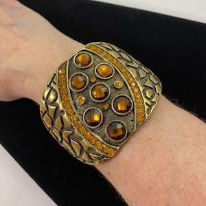 May include: A wide, gold-toned cuff bracelet with amber-colored gemstones. The bracelet features a textured, geometric design and is adorned with multiple round and oval-shaped amber stones. The cuff is worn on a wrist, showcasing its ornate details and warm color palette.