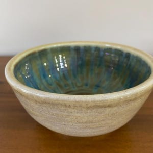 May include: A handmade ceramic bowl with a beige exterior and a blue and green interior. The bowl has a textured surface and a rounded shape, perfect for serving food or as a decorative piece. The bowl is approximately 6 inches in diameter.